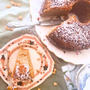 A Bundt cake on a white floral plate. There is a brown floral cake in the front what has once slice of the cake on it.