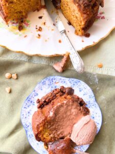 A Bundt cake in the background that has been cut. There is a slice of the cake on a blue floral plate. There is a scoop of chocolate ice cream on top.