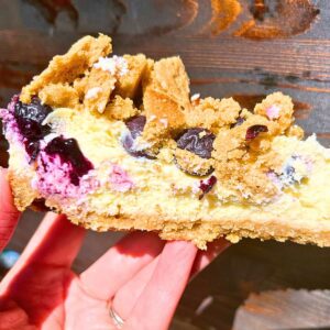 A woman holding up a slice of cheesecake with a blueberry and cookie crumble topping.