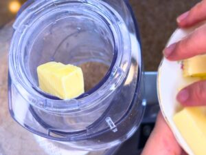 A woman adding cubes of butter through the funnel of a food processor.