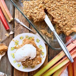 There is a cake in a glass casserole dish with a crumble topping. There is a knife in the casserole dish. There are some pieces missing. There is a slice of cake on a yellow floral plate. there is a scoop of vanilla ice cream on top. There are some pieces of rhubarb around on the table.