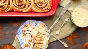 There is a cinnamon roll cut in half on a blue floral plate. There are more in a red casserole dish behind.