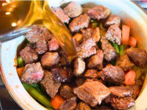 woman pouring beef stock into a pot with cubed beef and vegetables