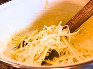 A white sauce pot with shredded cheese being added into it.