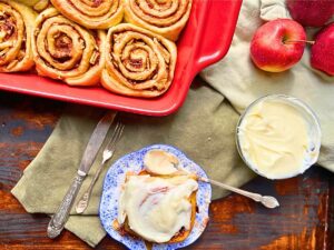 There is a red casserole dish with cinnamon buns inside. In front there is a blue floral plate with a cinnamon roll covered in white icing. 