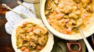 There is a pot with creamy beef stew inside. Beside is a white plate with a serving of the stew.