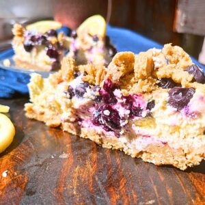 A slice of blueberry cheesecake with a cookie crumble topping. The slice is sitting on a dark wooden table.