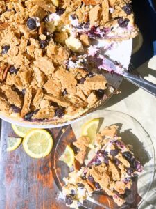 A cheesecake on a cake stand with a blueberry and cookie crumb topping. There is a slice of cake in front on a glass plate.