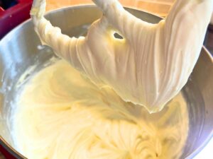 A metal mixing bowl with a whipped white cream cheese. There is a paddle attachment above with some of the whipped cream cheese mixture on it.