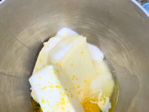 A metal mixing bowl with cream cheese, sugar, eggs, lemon zest and juice.