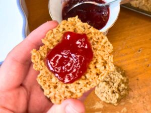 A woman holding an oatmeal cookie with jam spread in the centre.