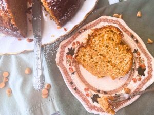 There is a slice of cake on a brown floral plate. In the background there is the whole cake.