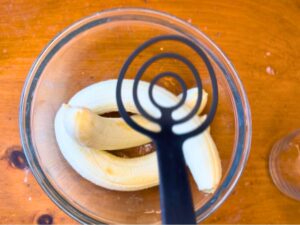 A glass bowl with 3 bananas inside. A woman is holding a masher.