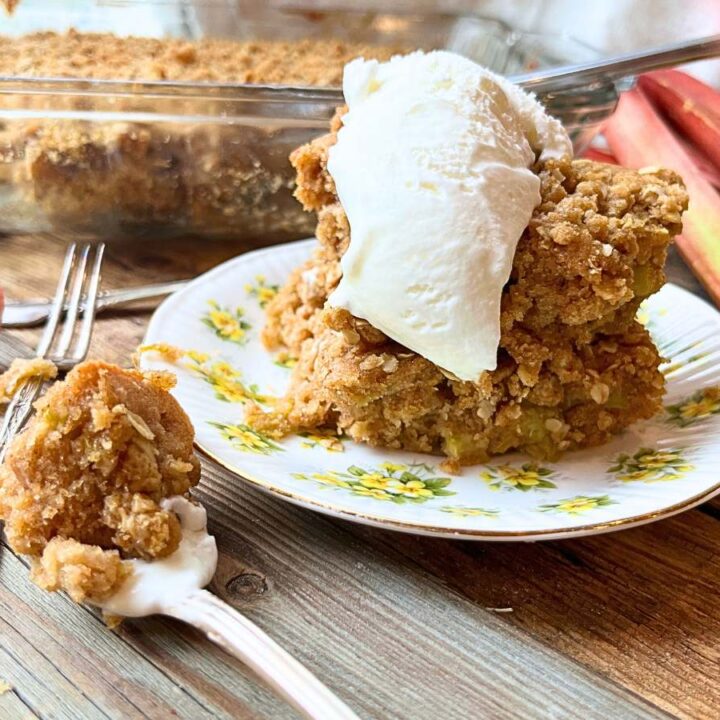 Rhubarb cake on a yellow floral plate with vanilla ice cream on top. In the background is more of the cake. There is a spoonful of cake beside.