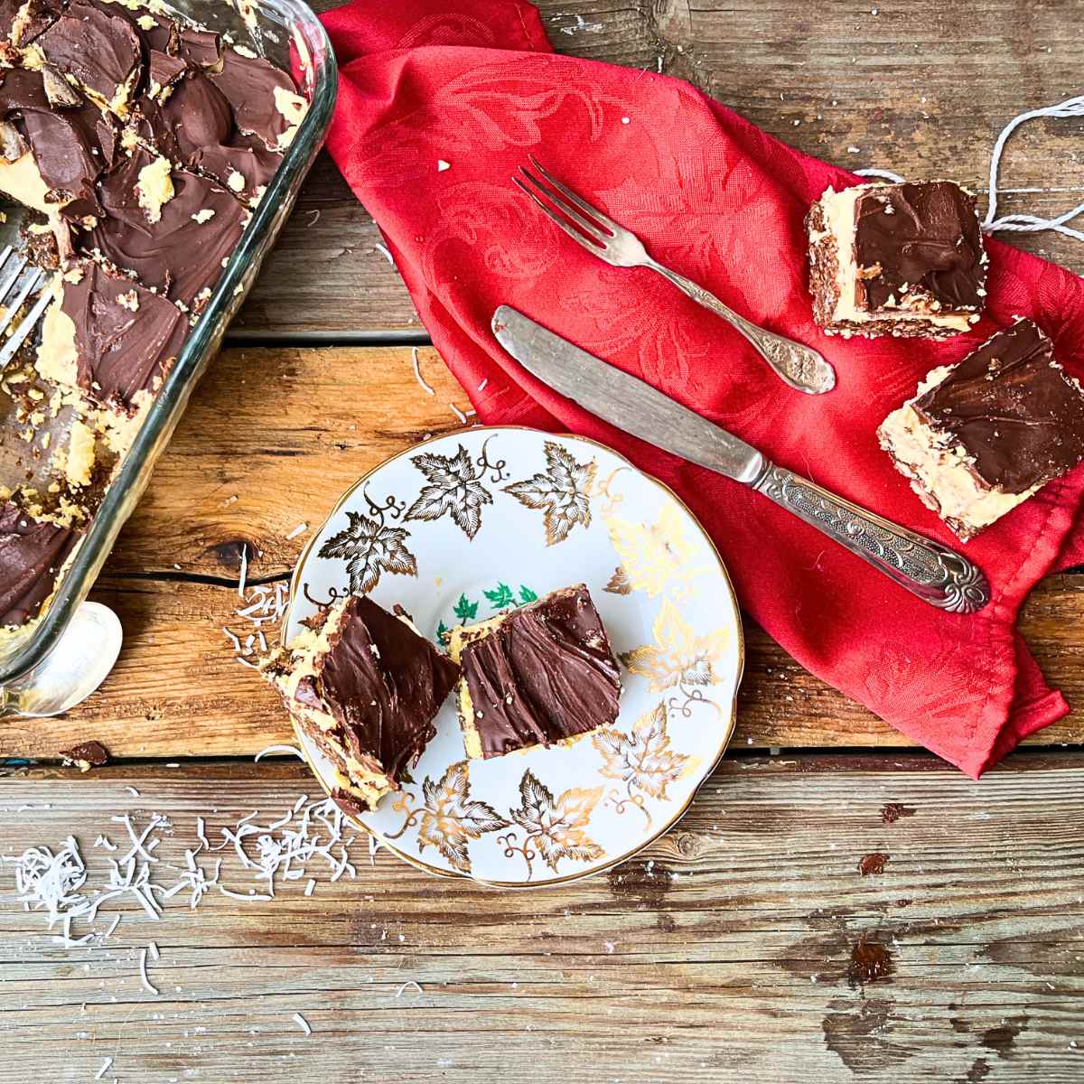 The top of wooden table. There is a gold maple leaf plate with 2 Nanaimo bars on it. There are two more on a red napkin.