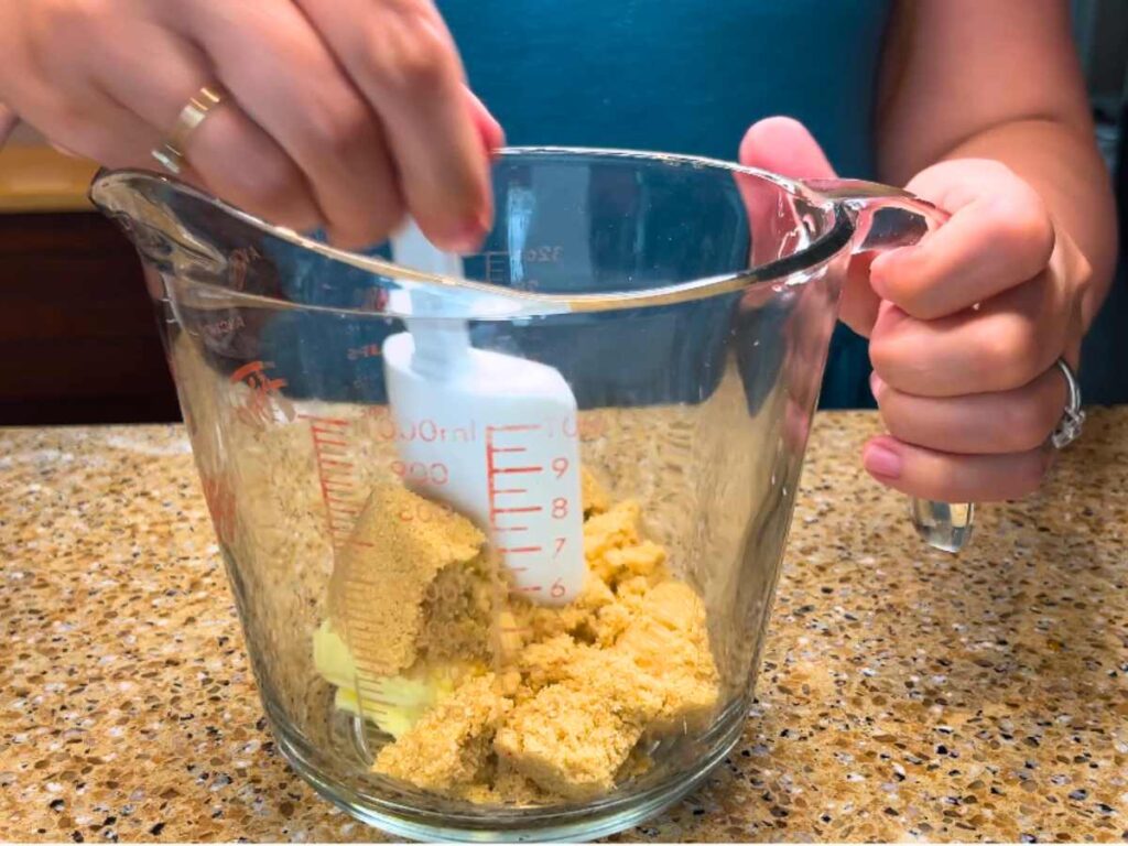 A woman mixing brown sugar and butter in a large glass measuring cup with a white spatula.