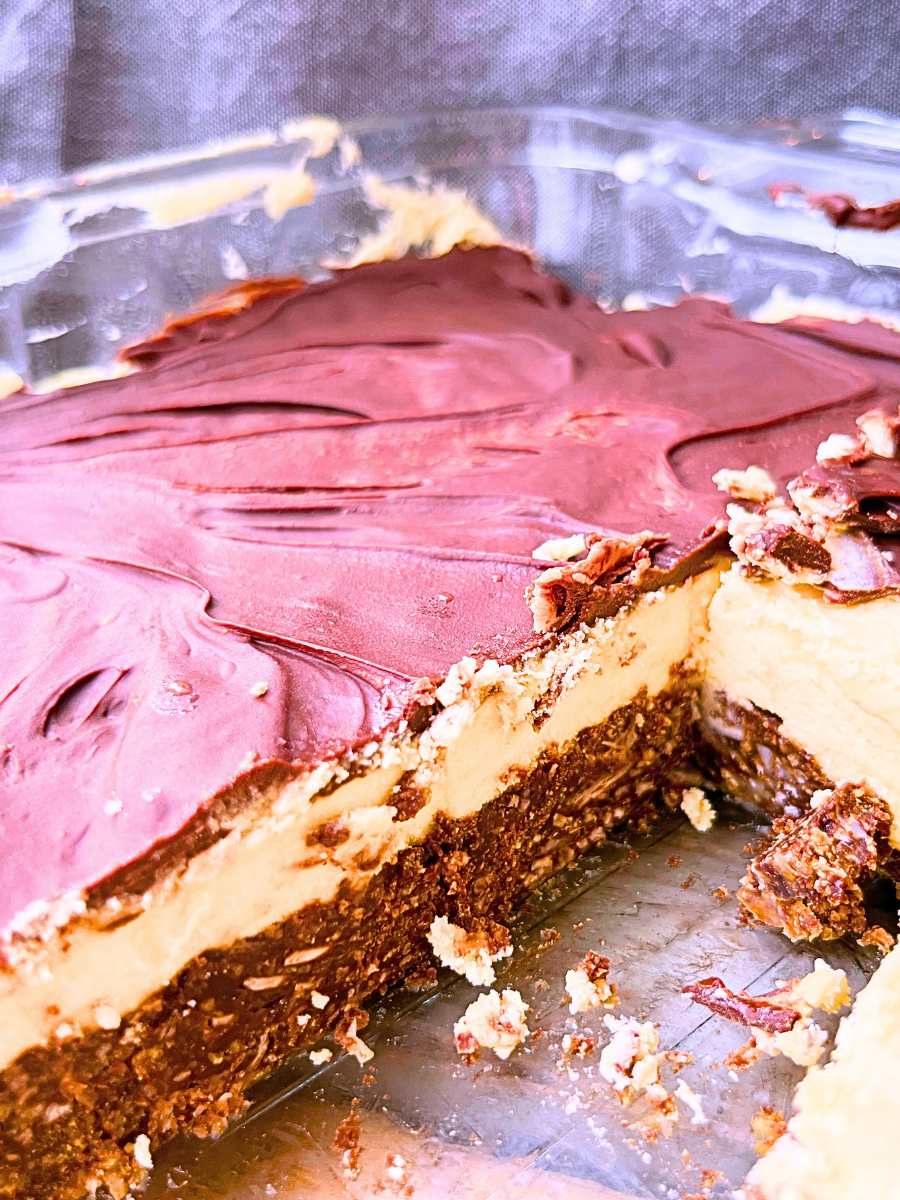 A square glass pan with Nanaimo bars layered in side. Some of the bars have been removed. You can see the three layers of the bars.
