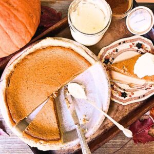 A sliced pumpkin pie. There is a slice on a brown floral plate. There is a pumpkin and jar of whipped cream in the background.