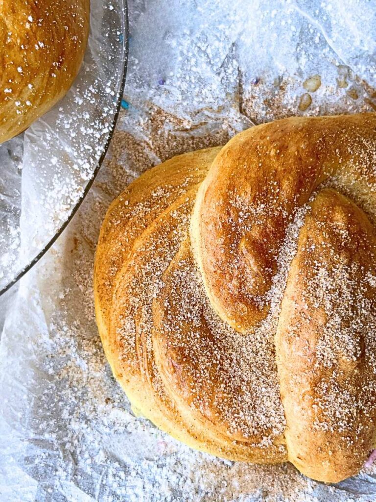 a sweet soft pretzel on a piece of parchment paper. There is another pretzel on a glass plate beside. The pretzel is covered in cinnamon sugar.