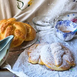 Two pretzels covered in powdered sugar. There are more pretzels on a glass plate in the background. There is a clue floral teacup and a green floral cream pitcher beside. 