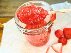 A jar of strawberry sauce. A woman is holding up a spoonful of sauce.