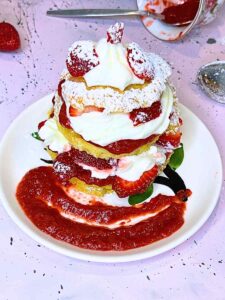 A strawberry shortcake with strawberry sauce on a small white plate. There are strawberries around and an overturned teacup with strawberry sauce in the background.