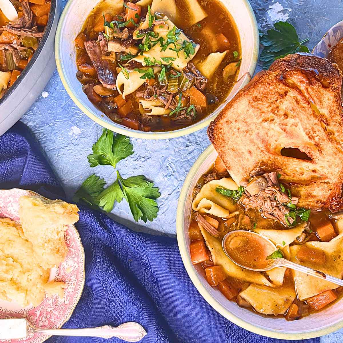 There is a blue table with two bowls of beef stew. There is a piece of toast dipped into the front bowl.