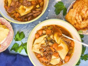 An overhead view of two bowls of beef stew on a blue table. There is a blue napkin in front and a piece of toast off to the side.