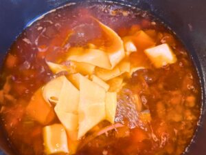 A pot of beef broth and vegetables with large noodles inside.