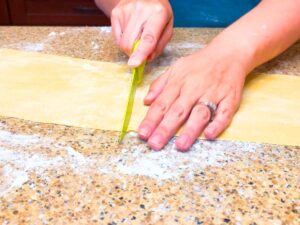 A woman cutting a long strip of pasta dough with a green knife.