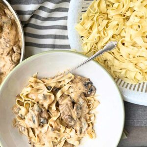 A bowl of beef and mushroom pasta. In the background is more of the sauce and more of the noodles in a colander.