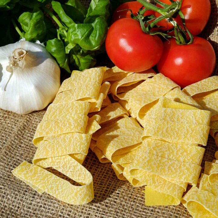 Fresh pasta noodles on a burlap fabric. There is basil, tomatoes, and garlic in the background.