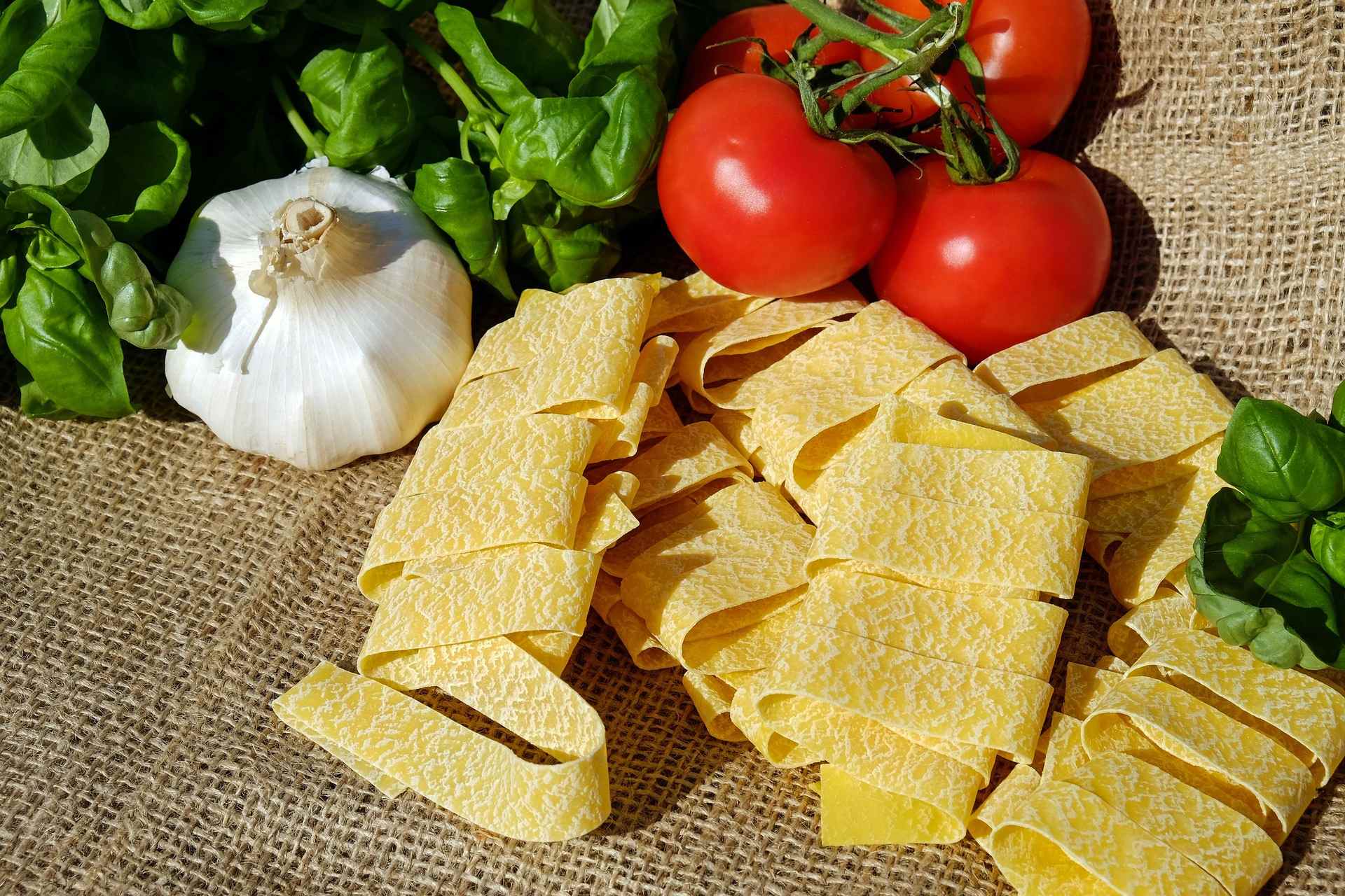 Fresh pasta noodles on a burlap fabric. There is basil, tomatoes, and garlic in the background.