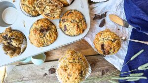 There are some chocolate chip muffins in a muffin tin. There are a few outside the tin and some pieces of chocolate around.