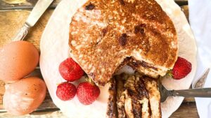 A stack of chocolate chip pancakes on a white plate. There are strawberries beside and a slice of pancakes cut out at the front.