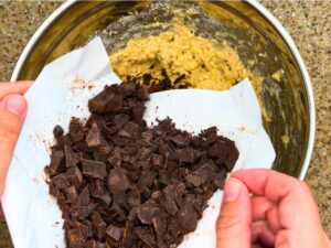 A woman pouring chocolate chunks into a bowl of muffin batter.