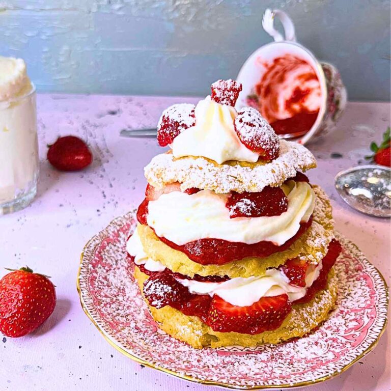A strawberry shortcake on a pink floral plate. There are whole strawberries around a overturned teacup with strawberry sauce in the background and a cup with whipped cream off to the side.