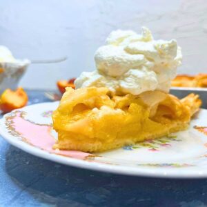 There is a slice of peach pie on a pink floral plate. There is more of the pie and peaches in the background.