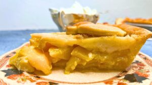 There is a side view of a peach pie with a cup of cream in the background.