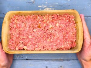 A woman holding a loaf pan with a raw meatloaf inside.