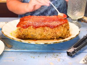 A woman spreading chili sauce onto a cooked meatloaf.