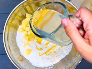 There is a woman pouring water and vinegar into a metal bowl with pie dough ingredients.