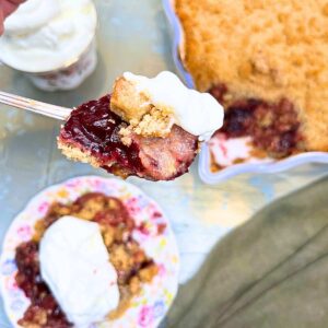 There is a woman holding a spoon full of cherry crumble with whipped cream on top. The rest of the cherry crumble is in the background.