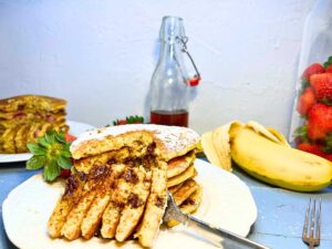 Pancakes that have a slice out of them. There is maple syrup, bananas, and strawberries in the background.