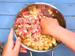 A mixture of ground meat, bread crumbs, and seasoning being mixed in a metal bowl.