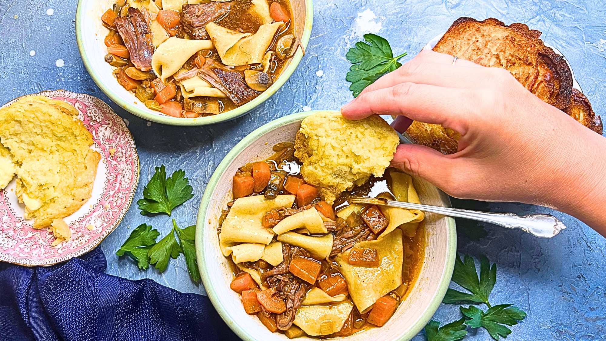 There is a woman dipping a biscuit into a beef stew. There is another bowl and more of the biscuit behind.