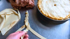 Someone is braiding a length of pie dough. There is an unbaked pie in the background.