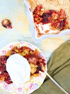 There is a serving of cherry crumble with a scoop of whipped cream on top. There is more of the crumble in the background.