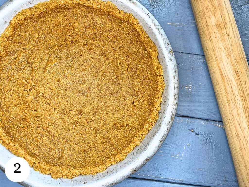 A graham cracker crust in a metal pie plate. There is a rolling pin beside.