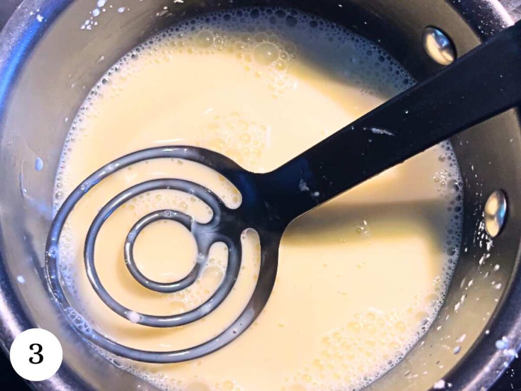 A small black sauce pot with milk inside. There is a whisk on top.